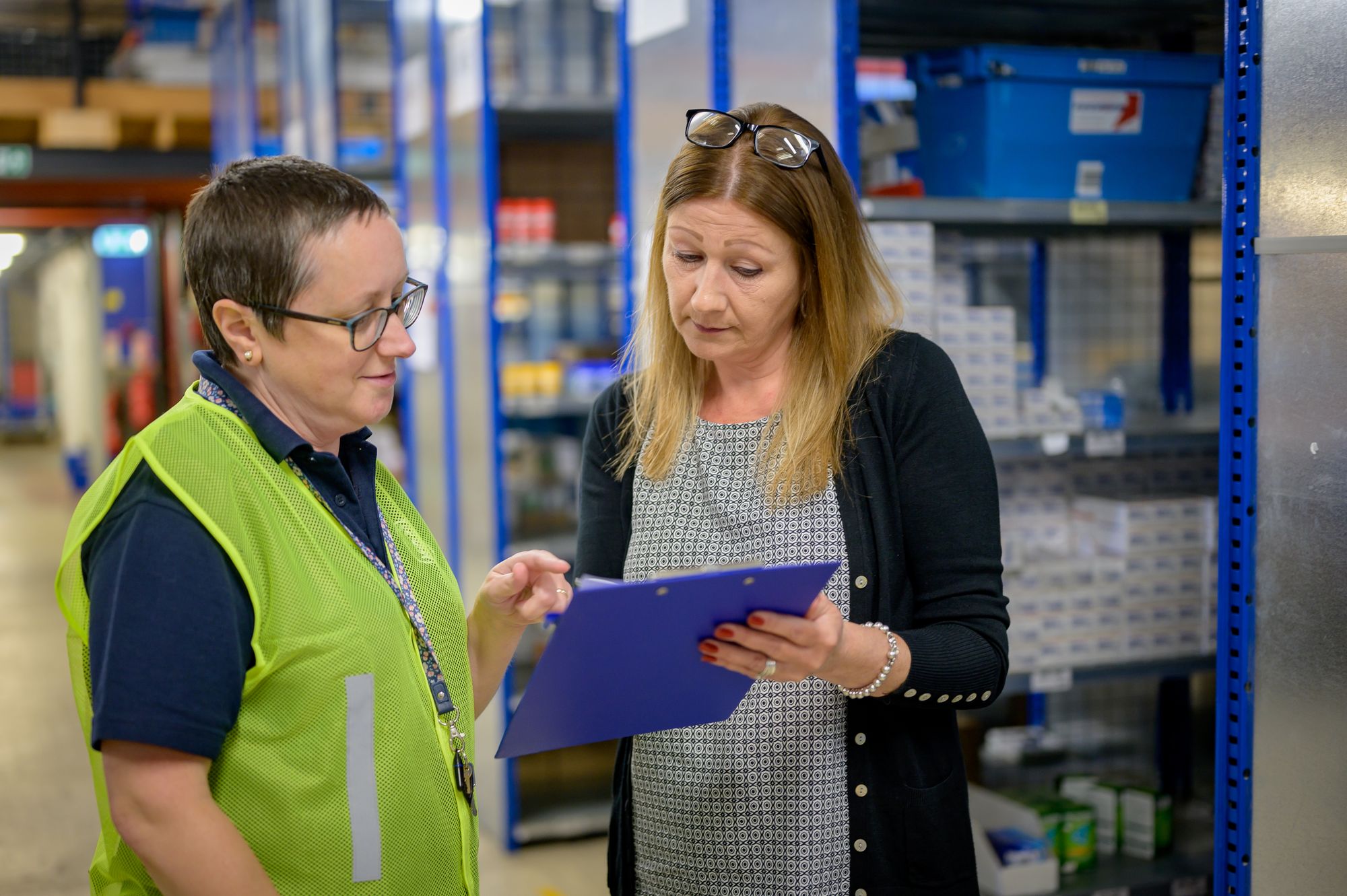 Two colleagues reviewing a stock checklist beside warehouse shelves